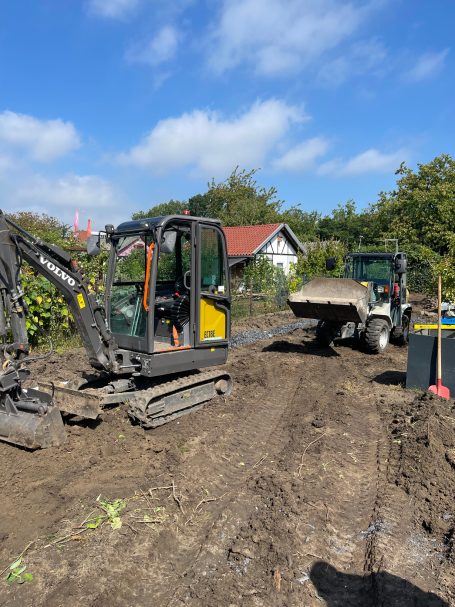 Bagger und Lkw auf einer Baustelle mit blauem Himmel und Bäumen im Hintergrund.
