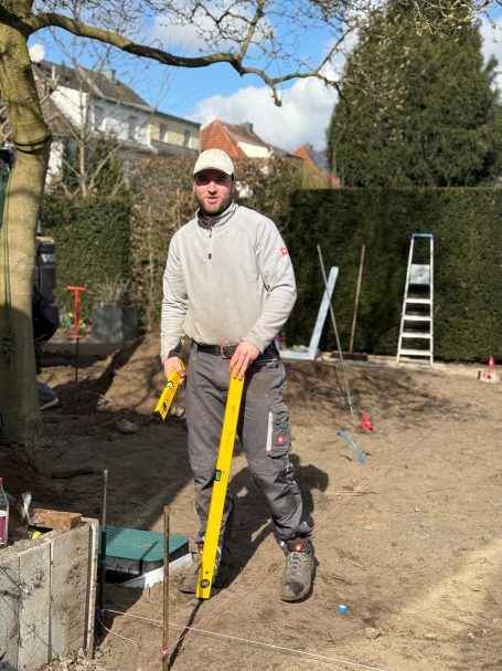 Mann mit gelbem Werkzeug steht auf einer Baustelle, umgeben von Werkzeugen und Bäumen.