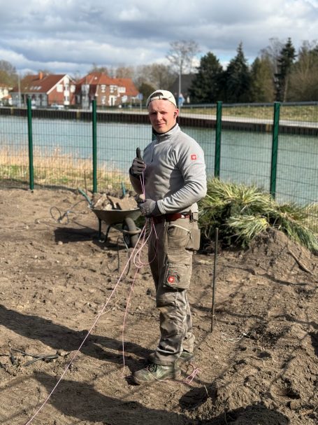 Mann mit Schaufel und Werkzeug steht auf unbefestigtem Boden am Wasser.
